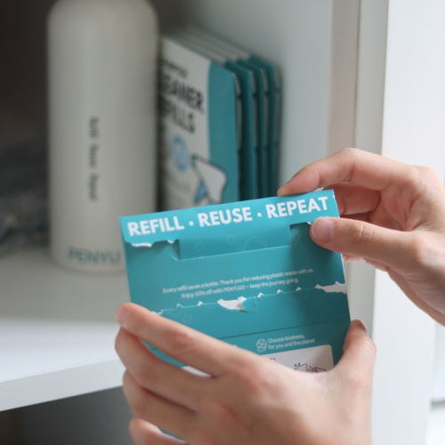 Person holding a blue refillable packaging with 'REFILL - REUSE - REPEAT' text, with a shelf in the background.
